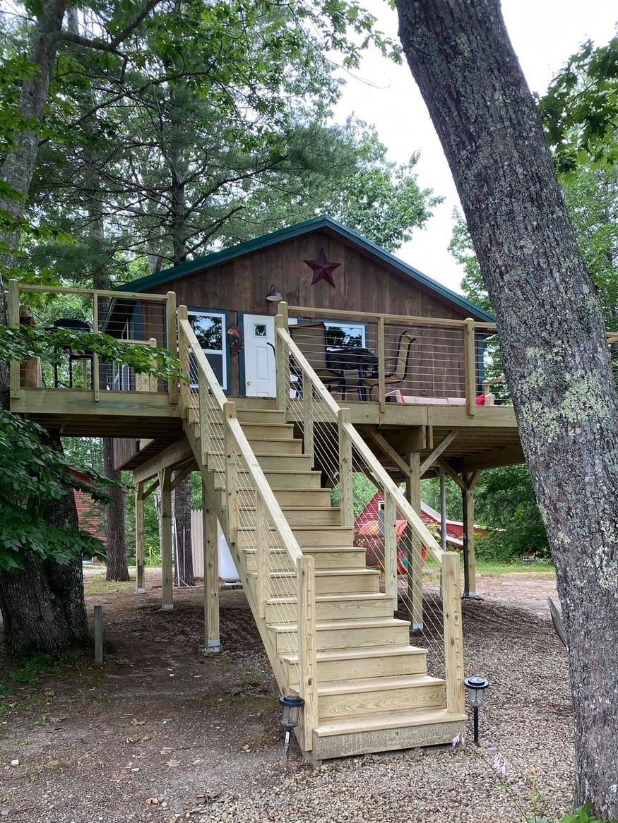 A wooden treehouse cabin is elevated on stilts, surrounded by trees. A staircase with wooden railings leads up to a porch that features seating options. The exterior is decorated with a star accent, enhancing the rustic charm of the setting.