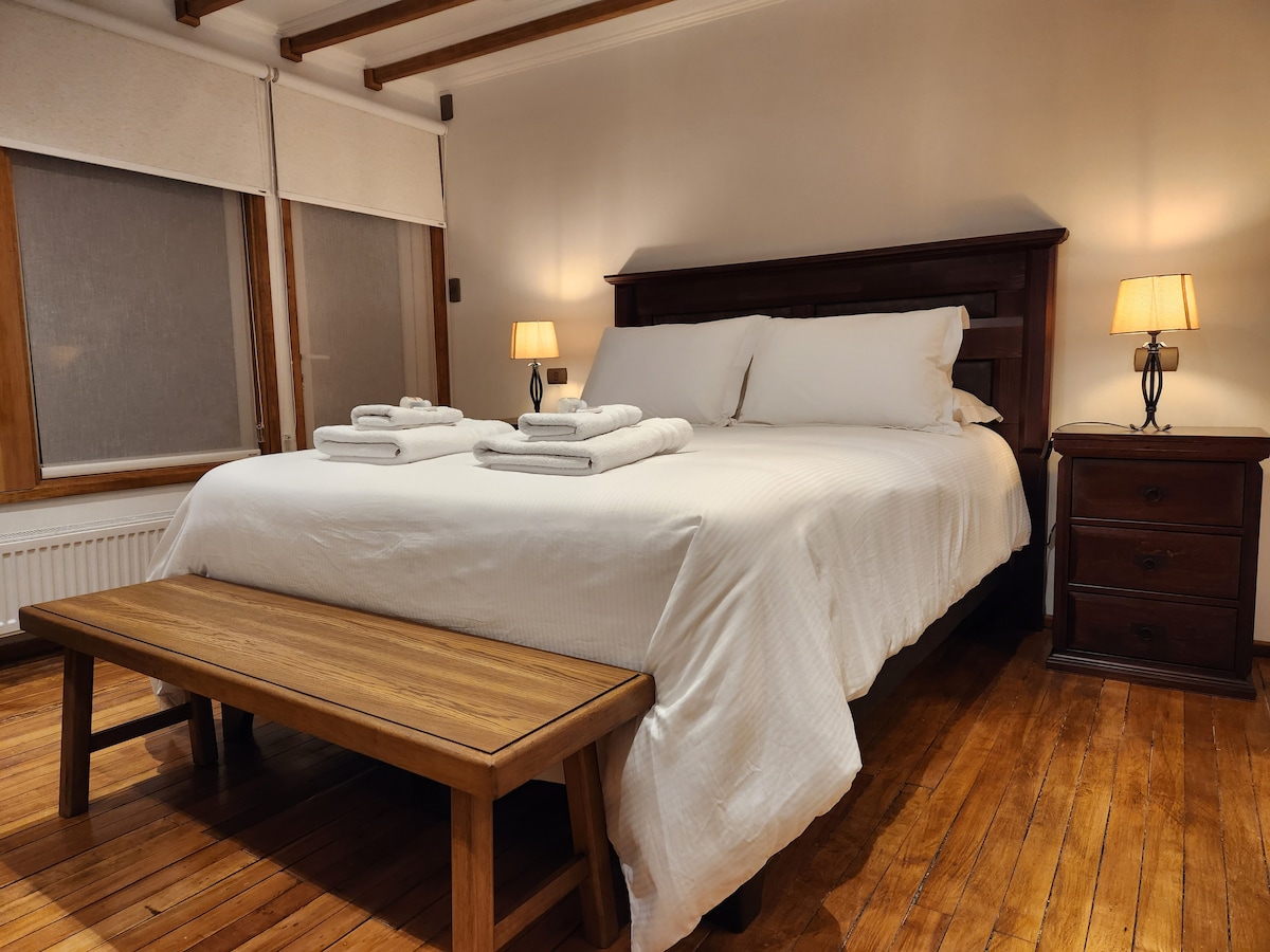 A warmly lit bedroom features a spacious bed with crisp white linens. On each side, bedside tables with soft-glow lamps provide gentle illumination. A wooden bench is placed at the foot of the bed, and natural light filters through partially covered windows.