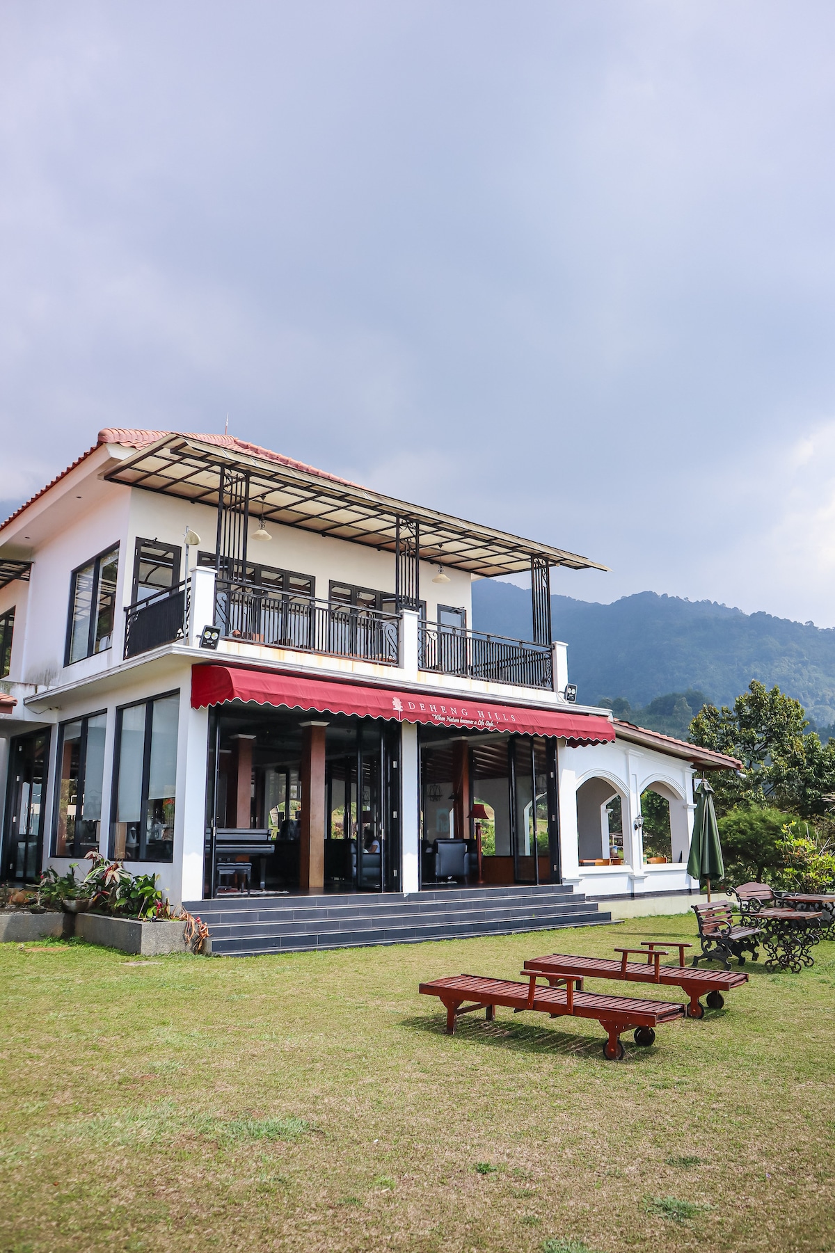 The villa's exterior showcases a modern design with large windows and a red awning. A spacious lawn features wooden seating areas surrounded by landscaped greenery. The backdrop consists of rolling hills and misty mountains, creating a serene outdoor setting.