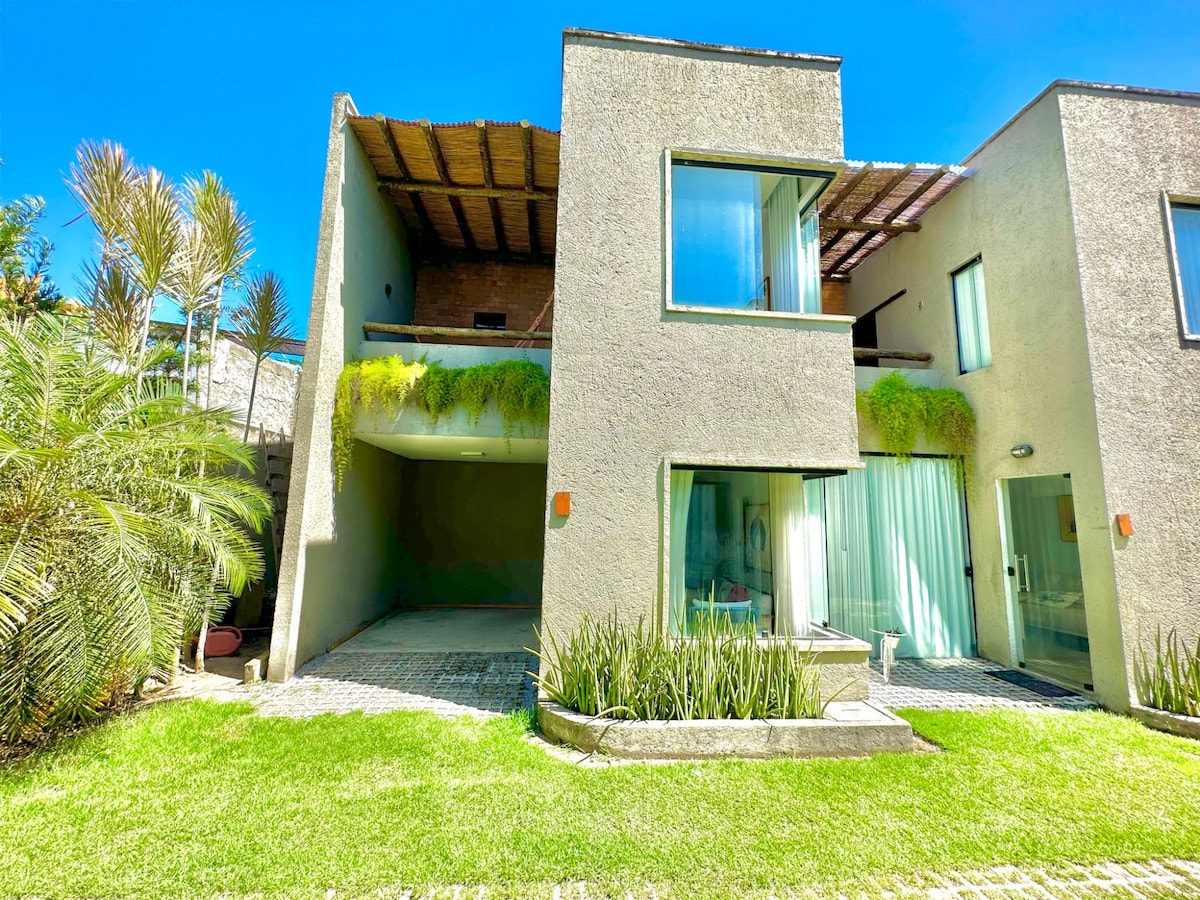The exterior of a modern two-story house is visible, featuring large glass windows that allow natural light to enter. Greenery adorns the building, enhancing the natural landscape. A spacious lawn surrounds the house, with a driveway leading up to the entrance.