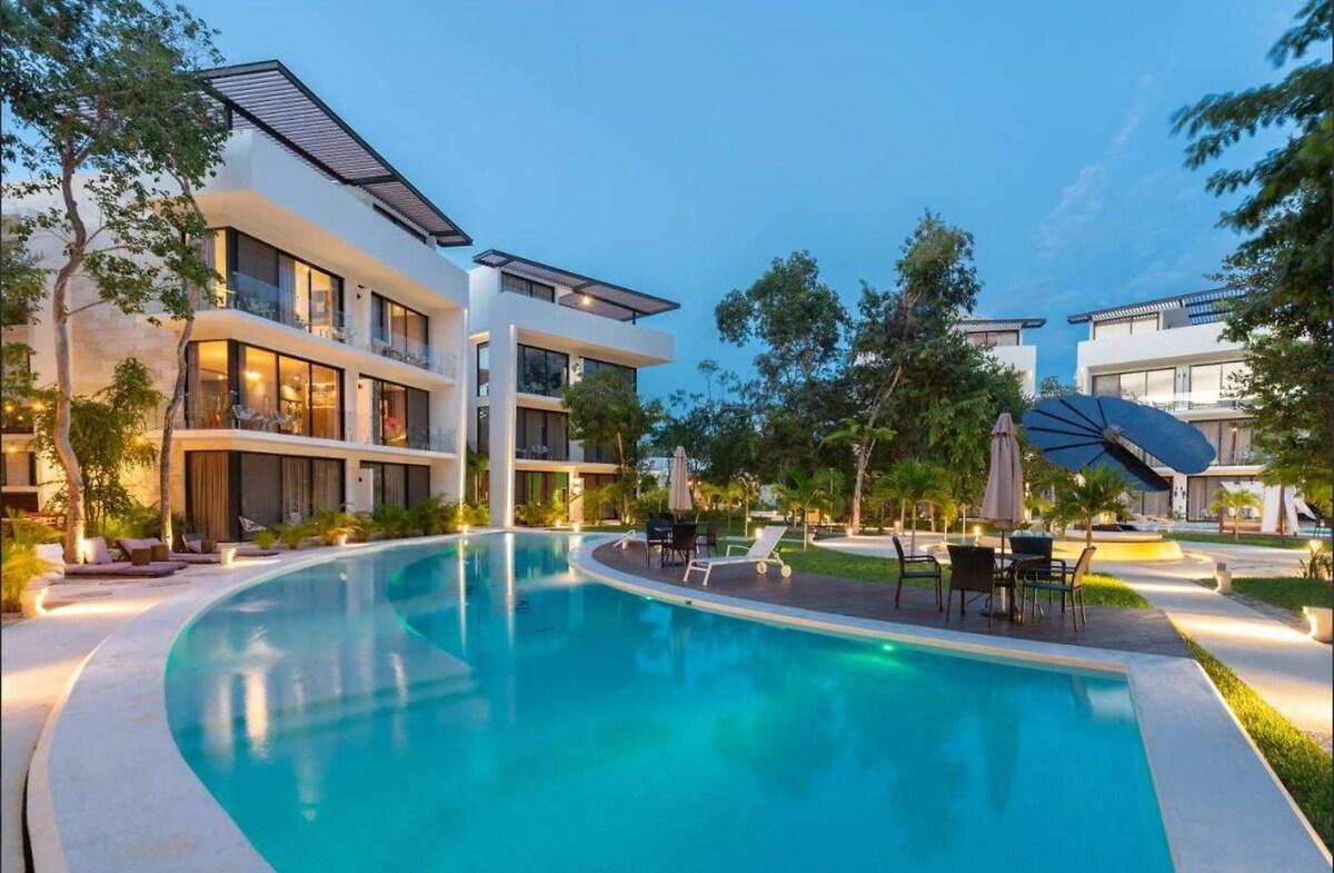 A serene communal area features a curving pool surrounded by lush greenery. Lounge chairs and umbrellas provide seating options, while nearby palm trees add to the tranquil setting. Modern buildings are seen in the background, highlighting the integration of natural elements with contemporary architecture.