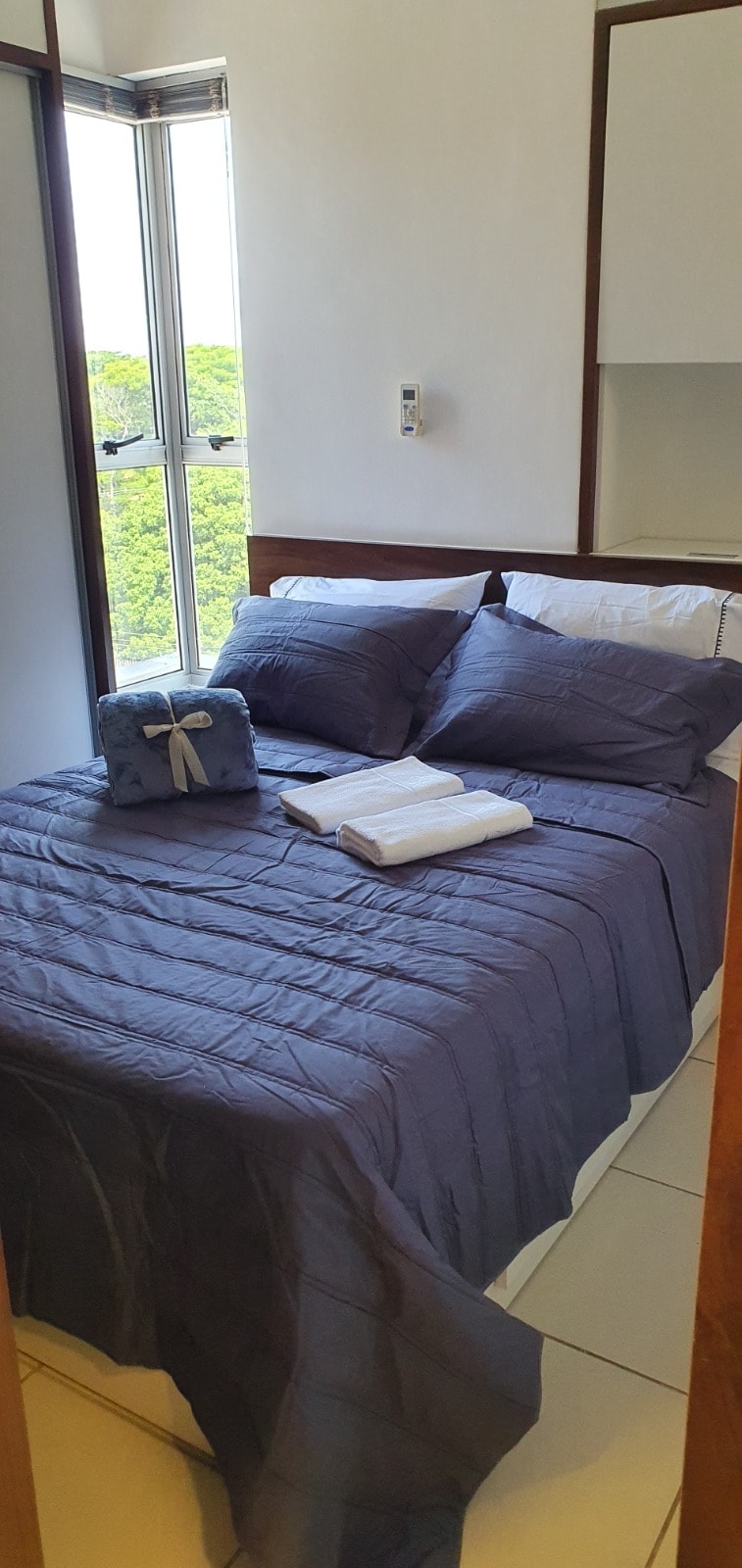 A sleeping area is presented with a spacious bed covered in a textured navy blue bedspread. Several pillows are arranged neatly against the headboard. A small towel is placed on the bed, alongside an additional folded towel, with a view of greenery visible through large windows.