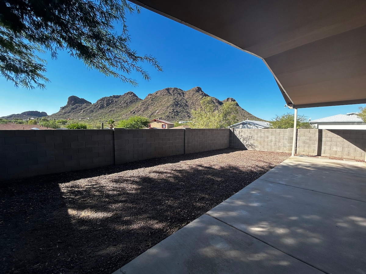 The outdoor area presents a gravel surface with a concrete patio, bordered by a low wall. The backdrop features distinct mountain peaks under a clear blue sky, enhancing the natural setting of the home.