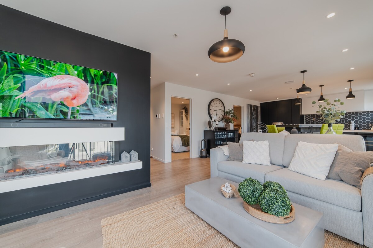 A modern living area is showcased, featuring a light-colored sofa adorned with decorative pillows. A sleek television screen displays vibrant imagery, while a wide, natural rug lays beneath a low coffee table, accented with greenery. The room benefits from ample natural light and open space.