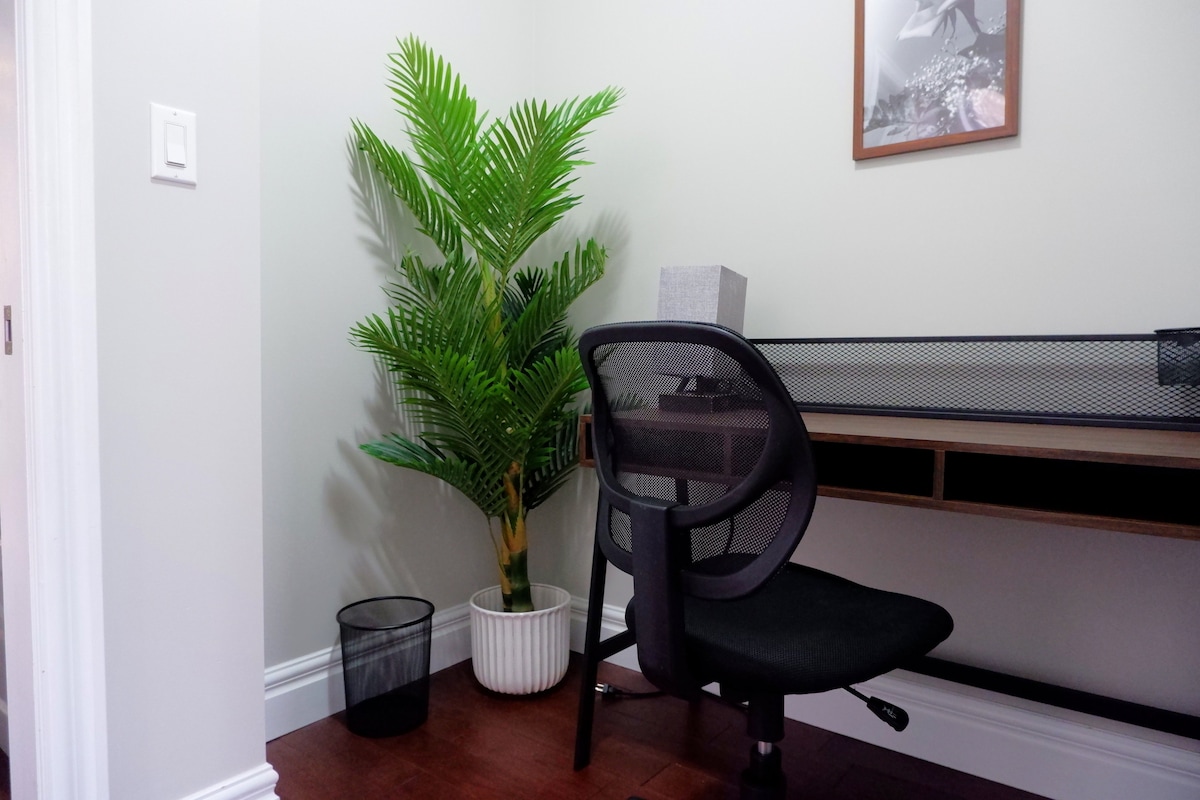 A small workspace is highlighted with a modern desk featuring a mesh-backed chair. A potted plant adds a touch of greenery beside a wastebasket. The walls are painted in a soft grey tone, providing a clean and comfortable environment.