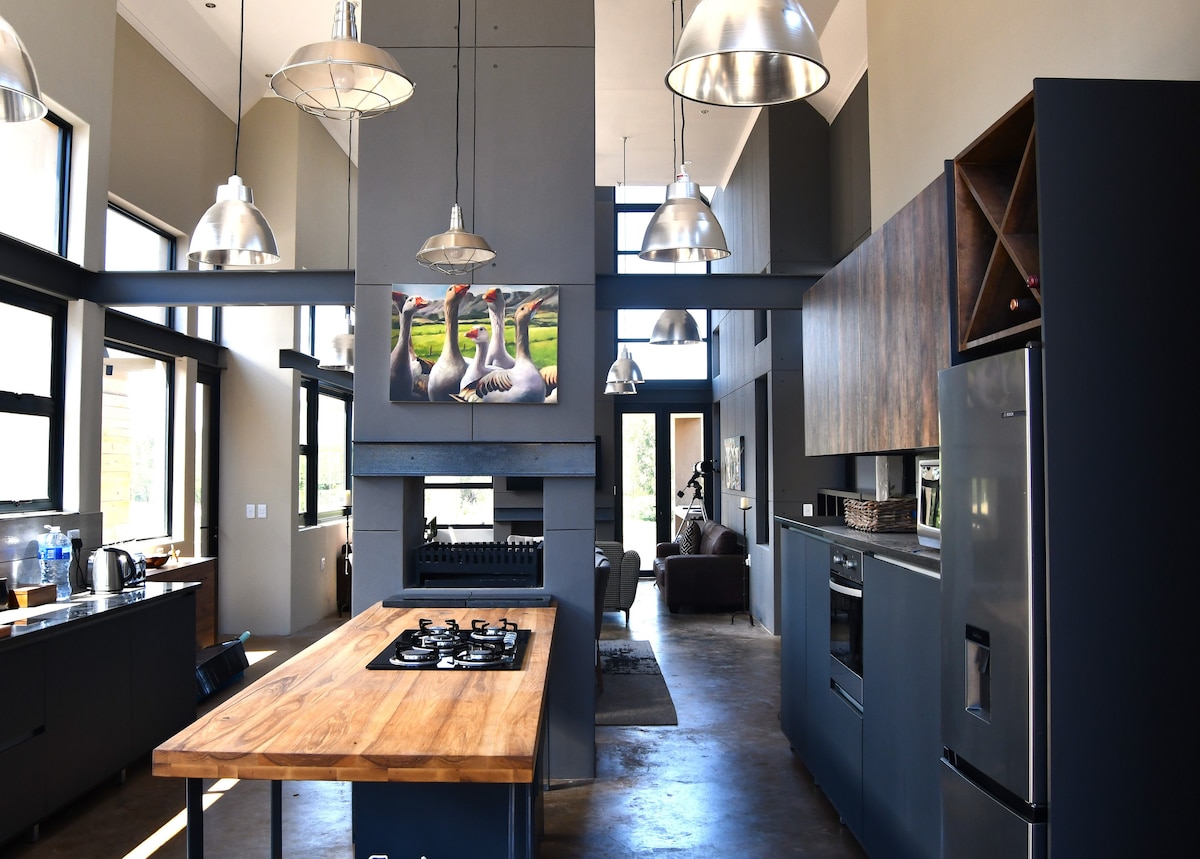 A modern kitchen is showcased, featuring an expansive wooden island with a gas cooktop. Open shelving and cabinetry provide ample storage. Large windows allow natural light to fill the space, highlighting stainless-steel appliances and a distinct wall art piece depicting serene wildlife.