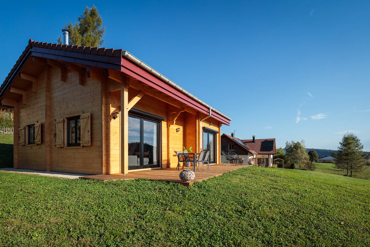 A wooden chalet features large glass doors opening onto a spacious deck, which overlooks a green landscape. The exterior is complemented by a sloped red roof and wooden accents, creating a harmonious blend with the natural surroundings.