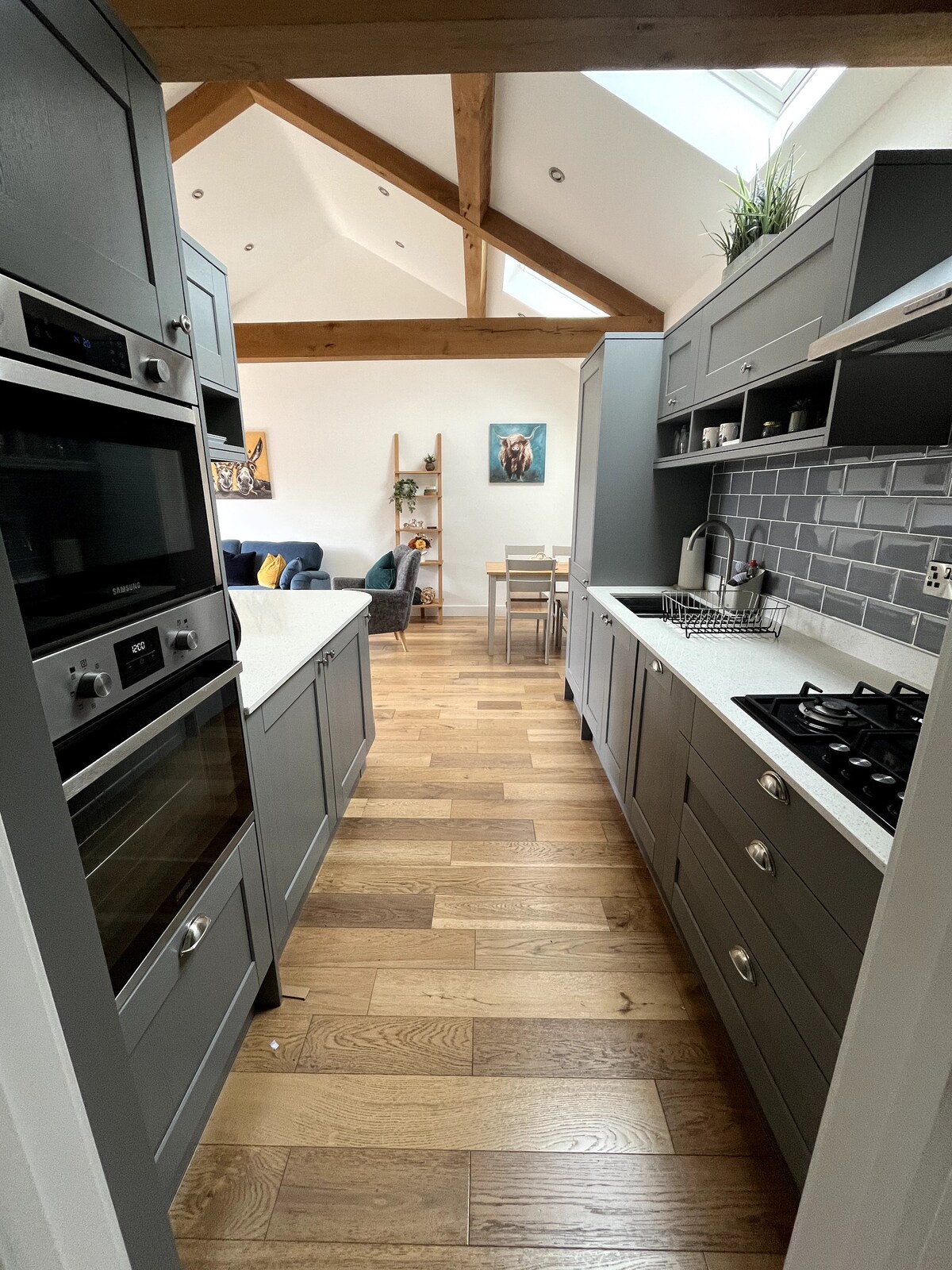 A modern kitchen is showcased with grey cabinetry and white countertops. Stainless steel appliances, including an oven and gas stove, are present. Natural light filters through skylights, illuminating the space. The open layout leads to a dining area with a simple wooden table and chairs.