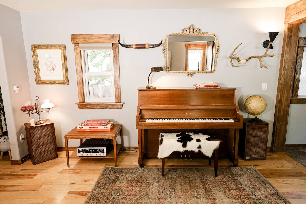 A charming music corner features a polished wooden piano equipped with a decorative cowhide bench. Framed art hangs on the walls alongside a mirror and unique décor pieces like a globe and antlers, adding character to the inviting wooden flooring.
