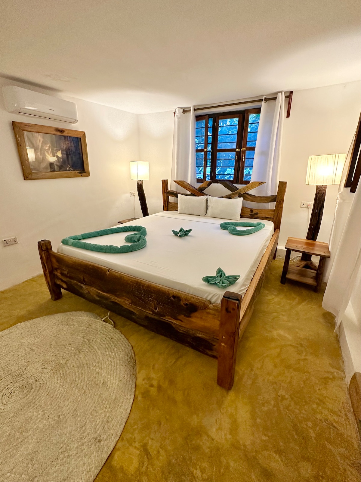 A cozy bedroom features a wooden bed adorned with decorative towels shaped like leaves. Two bedside lamps provide soft lighting, and a round rug is placed near the floor. A window with gridded panes allows natural light to fill the space.