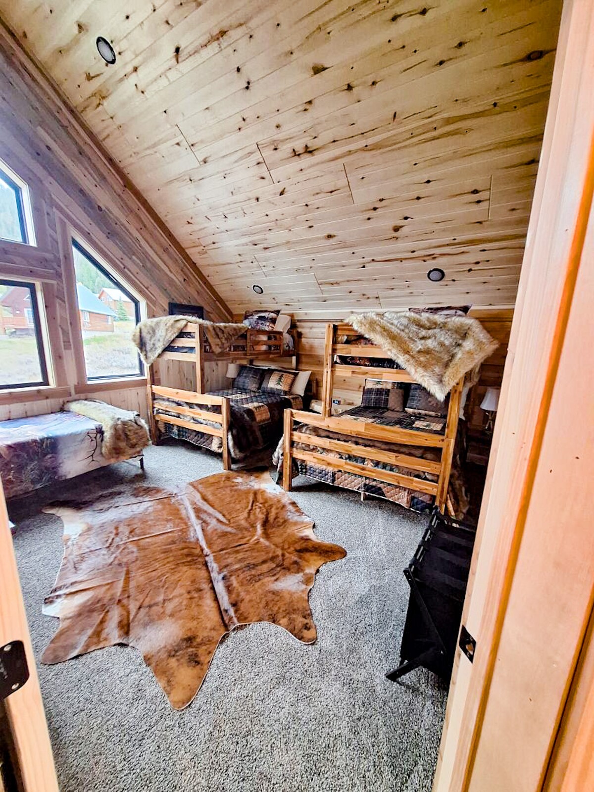 Een ruime slaapkamer in loftstijl, ontworpen voor groepen en gezinnen, met twee queensize bedden en drie eenpersoonsbedden. Grote ramen zorgen voor daglicht en uitzicht op de bergen, terwijl het interieur in hutstijl, knus beddengoed en een aan de muur gemonteerde tv.