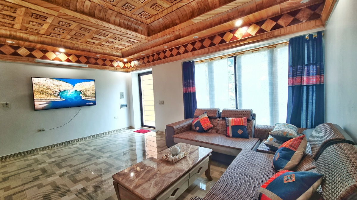 A spacious living area is presented, featuring a large television mounted on the wall opposite a comfortable seating arrangement. Natural light filters through large windows, complemented by patterned curtains. A coffee table with decorative items is centered on the tiled floor, enhancing the relaxed ambiance.