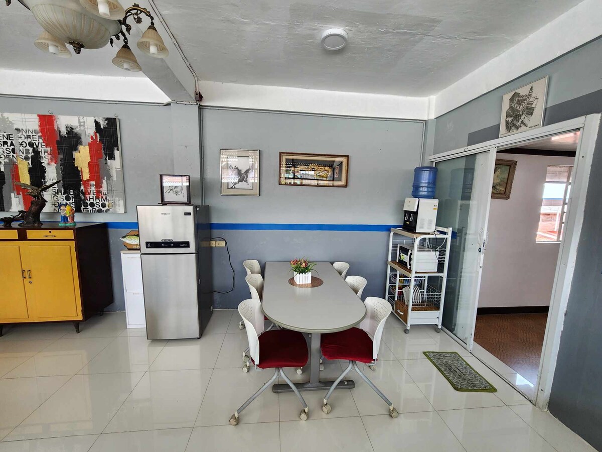 A dining area features a round table surrounded by four chairs, with a water dispenser in the background. A refrigerator stands next to a yellow cabinet, while artwork adorns the walls. Natural light emphasizes the glossy tiled floor and the overall spaciousness.