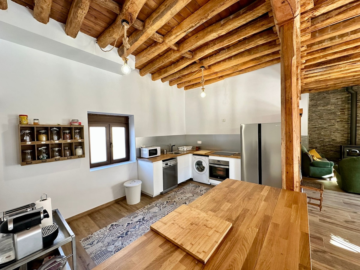 A well-appointed kitchen features rustic wooden beams overhead and modern appliances, including a refrigerator and washing machine. A wooden countertop provides ample workspace, while a small window allows natural light to illuminate the area. Open shelving displays jars, enhancing the functional design.