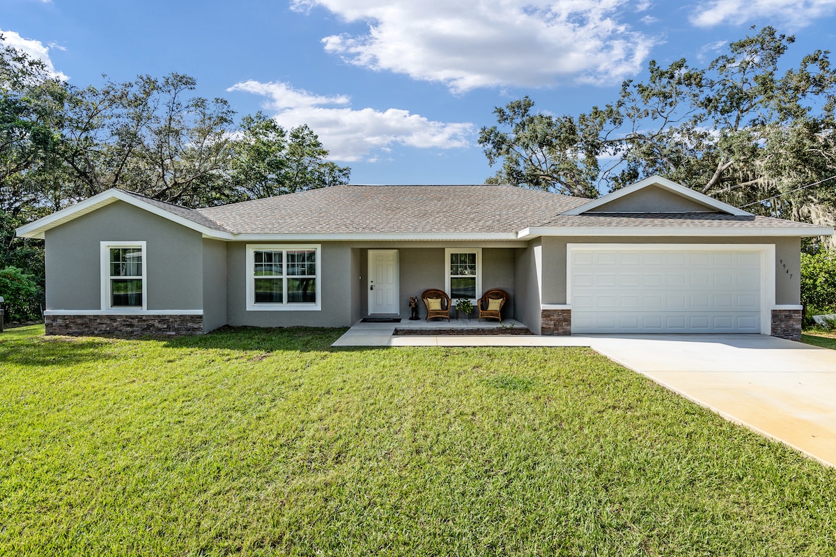 A newly constructed single-story residence is presented, featuring a welcoming covered porch with two chairs. The front yard is adorned with lush green grass, and the home is framed by tall trees under a partly cloudy sky.
