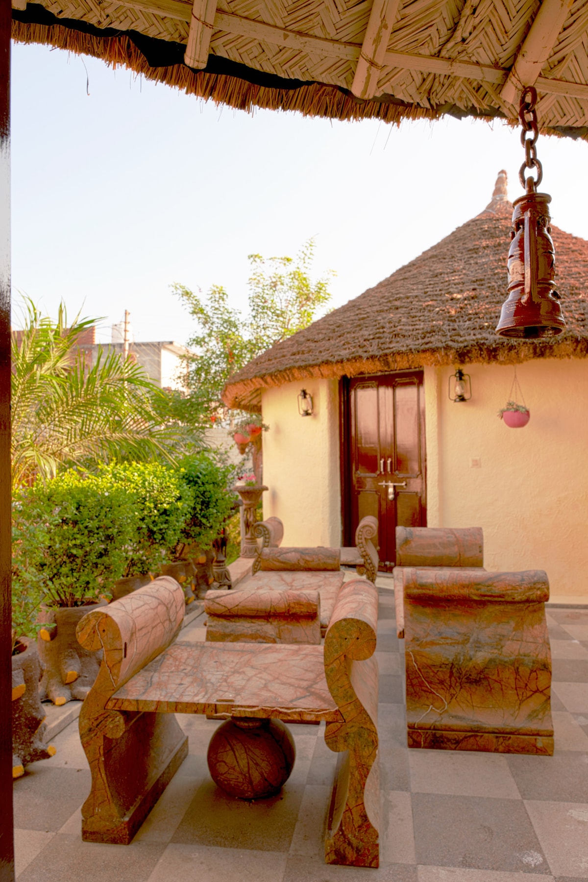 An outdoor seating area features rustic wooden furniture arranged around a stone table. Surrounded by greenery, the space is sheltered by a thatched roof, with a traditional lantern hanging from above, creating a calm and inviting environment.