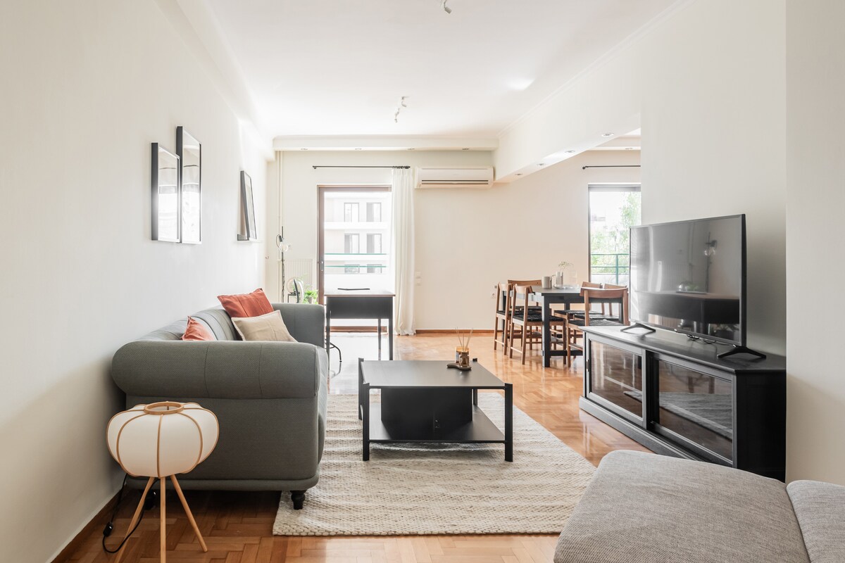 A spacious living area is designed with a comfortable gray sofa and a round side table. A large flat-screen TV is situated on a stylish media unit. Natural light fills the room through wide windows, with a dining table set nearby, enhancing the inviting environment.