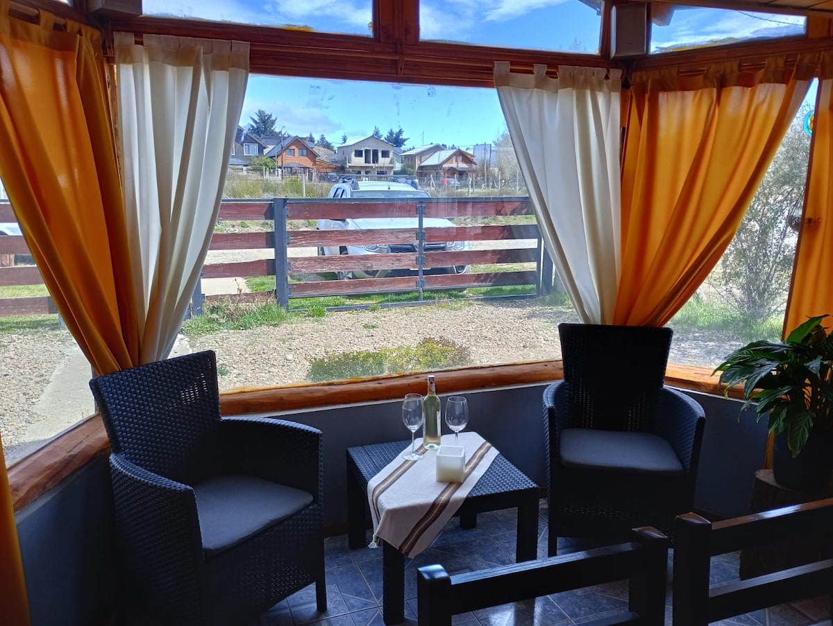 A sunlit seating area features two woven chairs facing a window. Light curtains in orange and cream frame the view of the landscaped exterior and nearby houses. A small table holds two glasses and a decorative item, inviting relaxed moments.