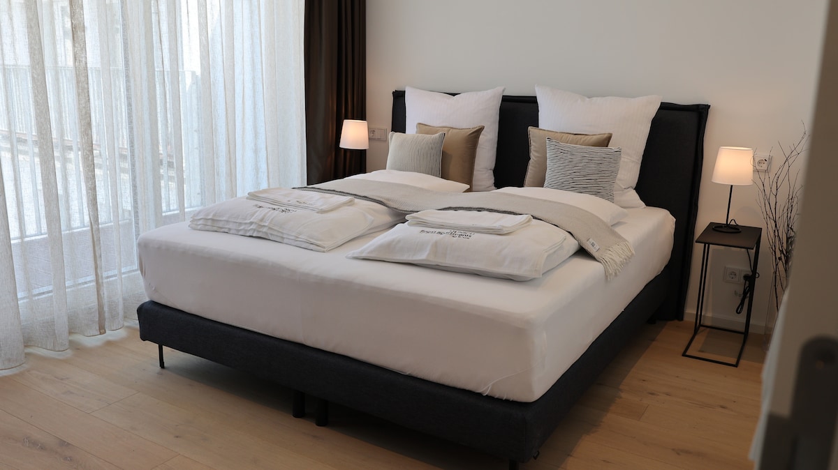 A spacious bedroom features a large bed with neatly arranged linens and decorative pillows. Soft lighting from bedside lamps enhances the calming atmosphere. Tall windows adorned with sheer curtains allow natural light to fill the room, creating a bright and airy environment.