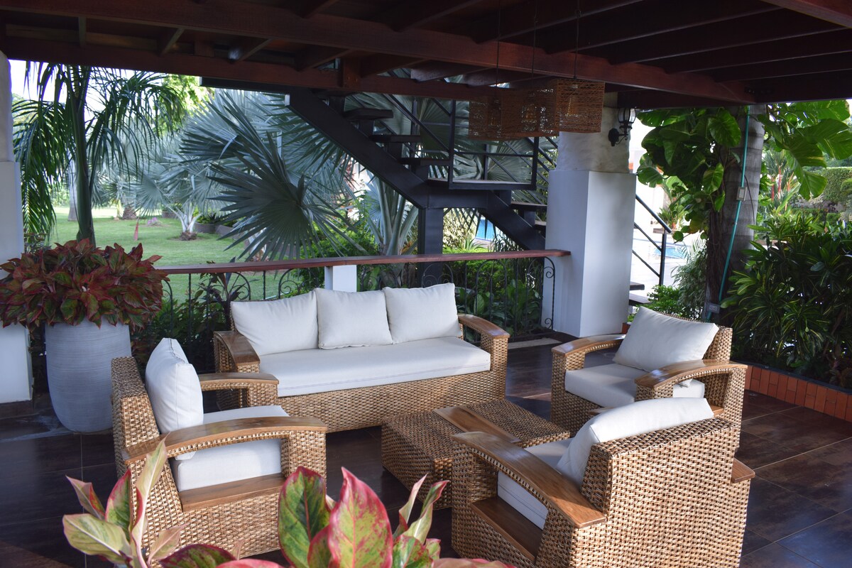 An outdoor seating area features a comfortable arrangement of woven furniture, including a spacious sofa and several armchairs. Lush greenery surrounds the space, with tall plants and palm trees providing a natural backdrop, inviting relaxation in a serene atmosphere.