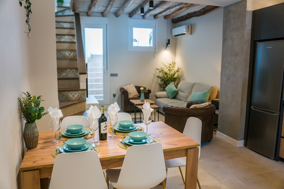 A modern dining area with a light wooden table set for four is visible, featuring dishes and cutlery. In the background, a comfortable sofa is arranged near a staircase leading to the upper level. Natural light streams in through a glass door, complemented by indoor plants.