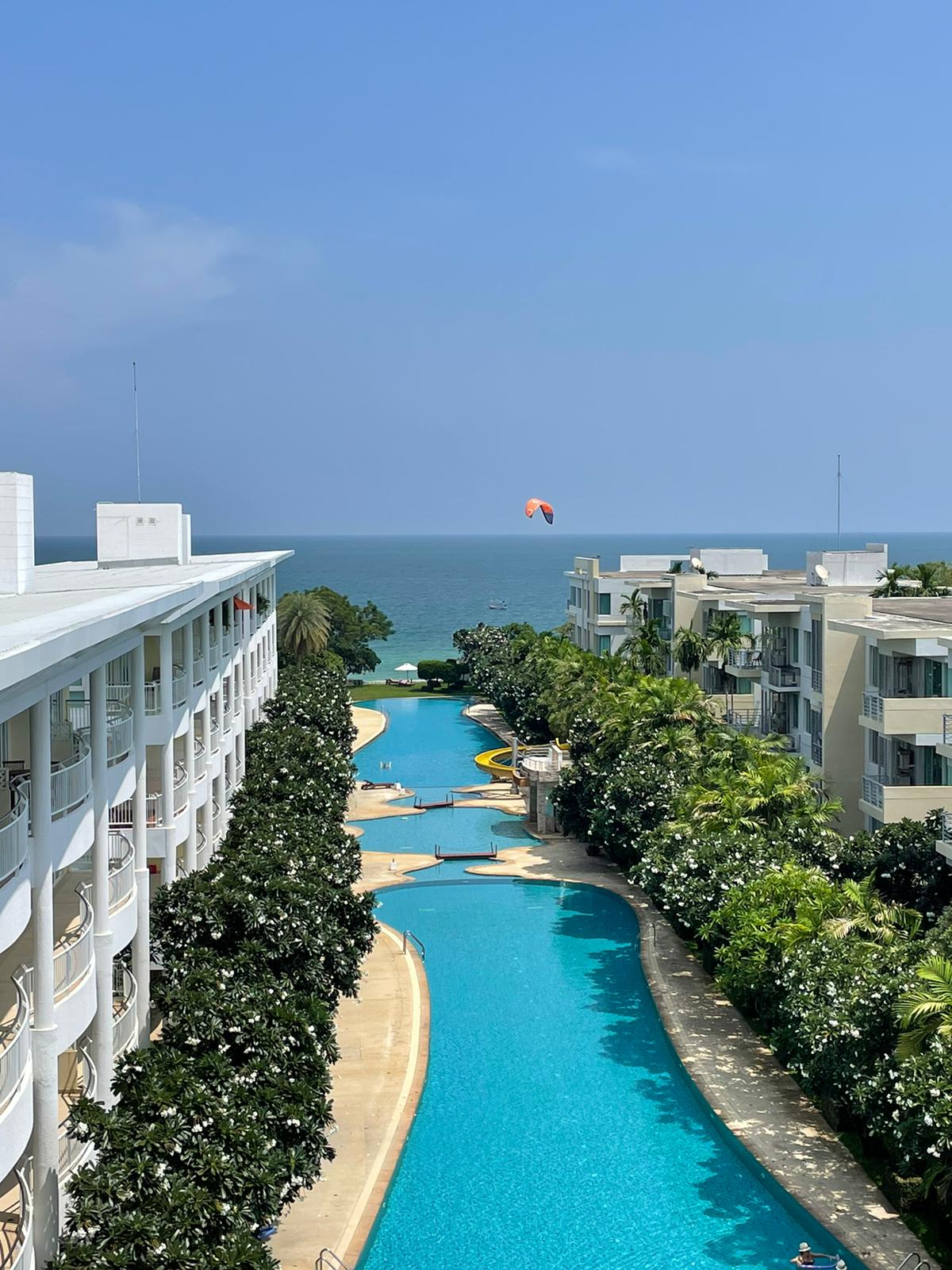 The image showcases a view of two elongated pools winding through a manicured landscape, leading towards the sea. Lush greenery lines the pools, and the ocean can be seen in the background, along with a brightly colored parachute in the distance.