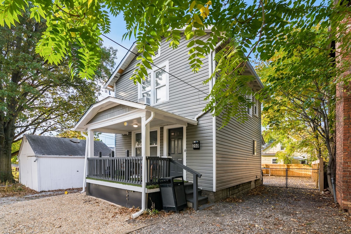 The exterior of the house features a light grey façade with white trim and a welcoming front porch, complemented by a protective overhang. Surrounding greenery adds a natural touch, while a gravel pathway leads to the entrance. A fenced yard extends behind the home.