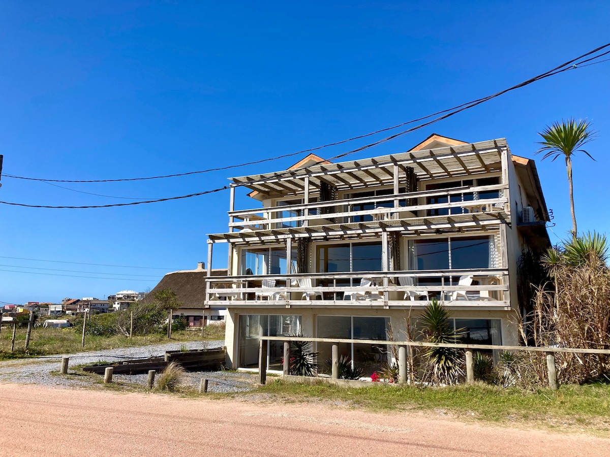 A modern building is presented with multiple balcony spaces, showcasing outdoor seating arrangements. The structure features large glass windows, allowing natural light to enter. Surrounding greenery and open skies contribute to a peaceful ambiance, while the proximity to the road provides accessibility.