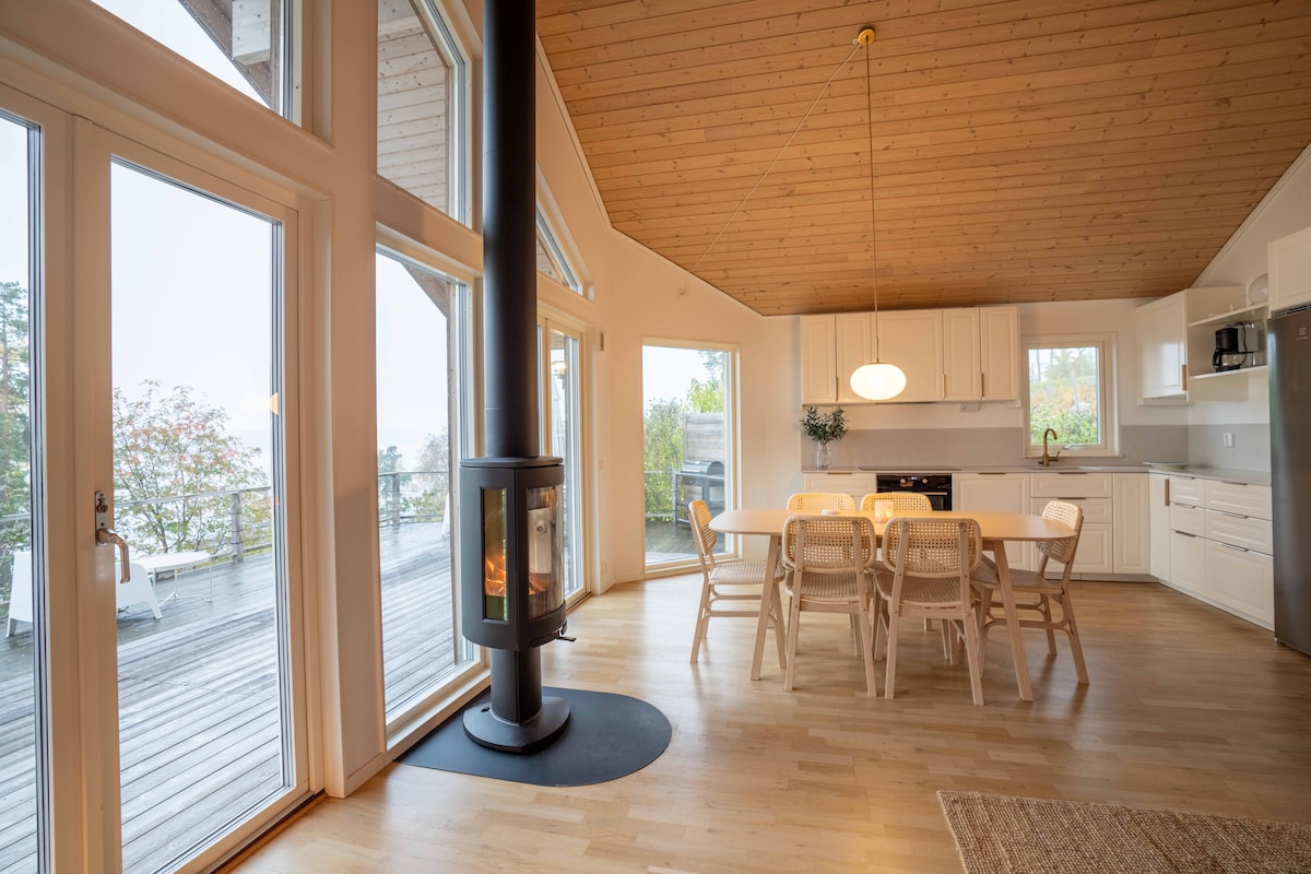 A spacious dining area features a sleek white table surrounded by light-colored chairs, complemented by a modern pendant light overhead. Large glass doors allow for natural light, with a cozy wood-burning stove positioned nearby, enhancing the room's warmth.