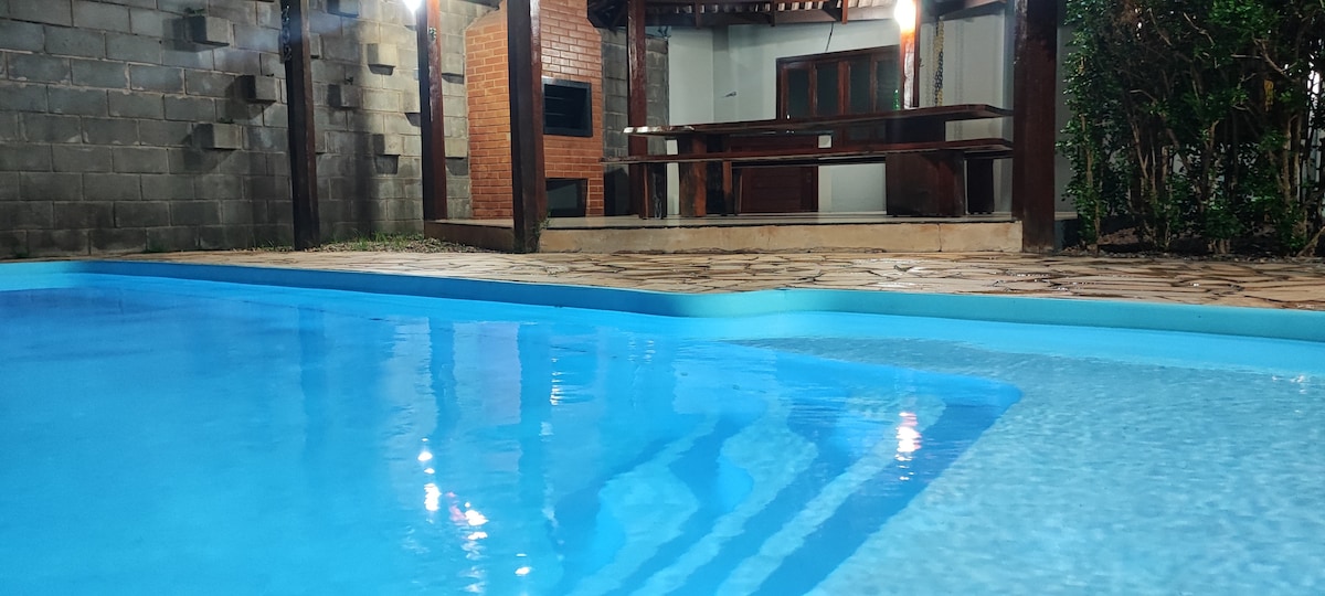 A clear view of a tranquil swimming pool reflects soft lighting from an adjacent outdoor kitchen area. The stone patio surrounds the pool, providing a spacious area for relaxation. Decorative greenery frames the space, enhancing the overall ambiance.