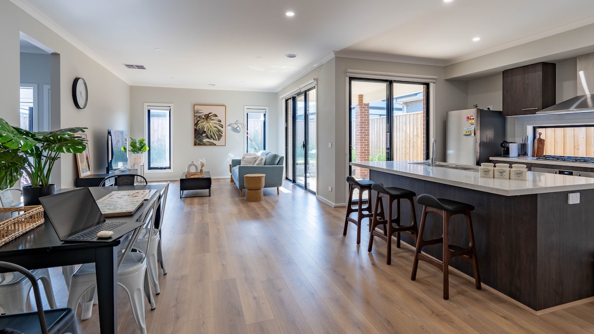 An open-plan dining and living area features large windows allowing natural light to fill the space. Stylish furniture is arranged around a sleek kitchen island, complemented by modern appliances and a dining table set for gatherings. A cozy seating area invites relaxation.