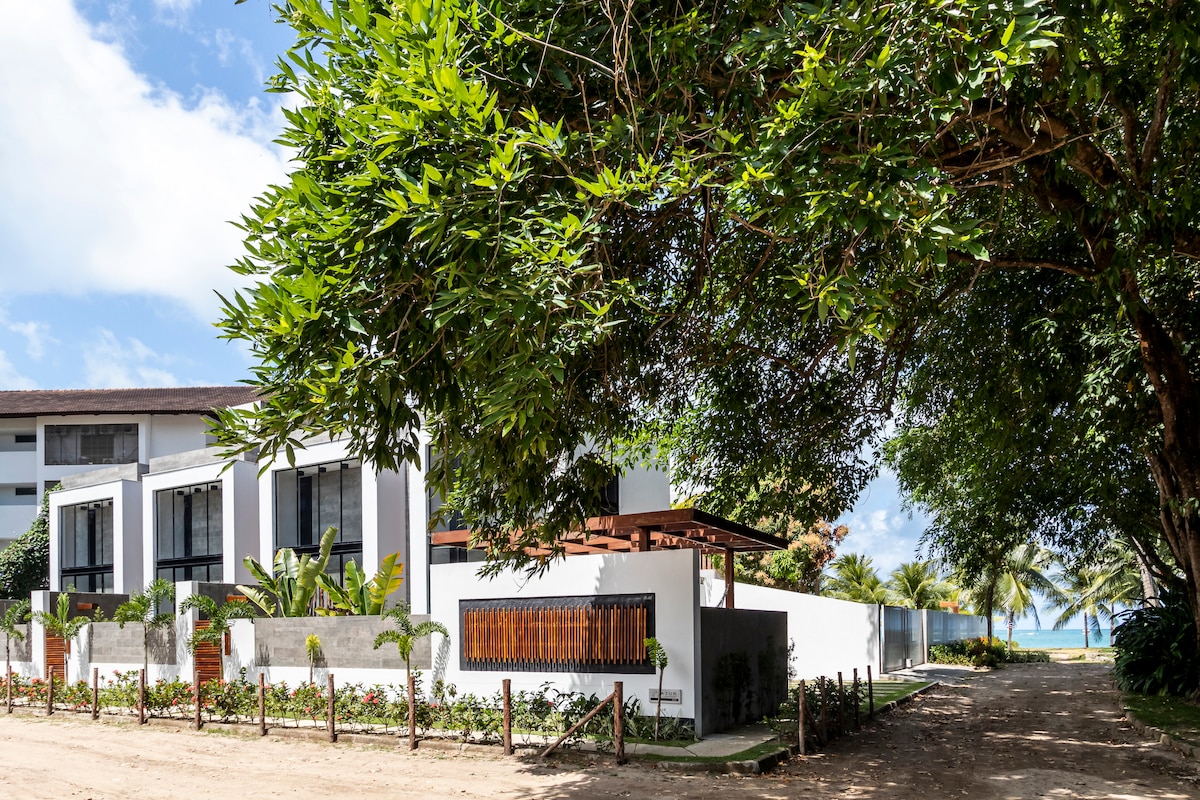 A modern house is captured from an angle that highlights its clean lines and expansive windows. Lush greenery surrounds the property, with palm trees visible in the background. The beach can be seen at a distance, emphasizing the proximity to the water.