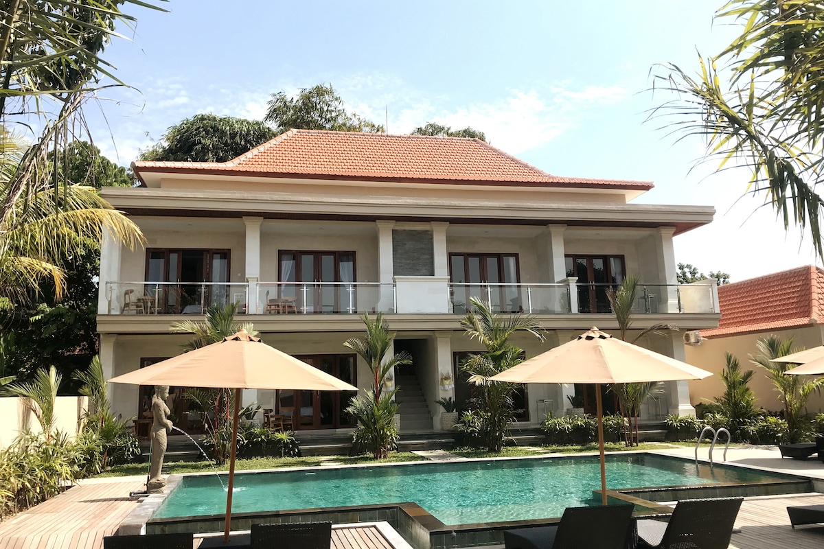 A modern villa features a spacious two-story structure with a red-tiled roof. Large glass doors open to a landscaped area with a swimming pool surrounded by tropical plants. Sun loungers and umbrellas provide shaded seating along the pool deck.