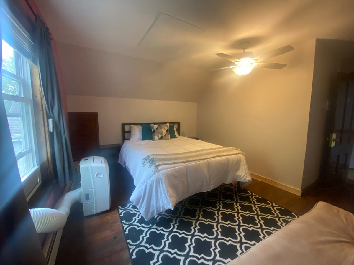 A spacious sleeping area features a soft bed covered with light-colored linens and decorative pillows. A fan is mounted on the ceiling, providing airflow. A large window allows natural light to enter, while a patterned rug adds texture to the floor.