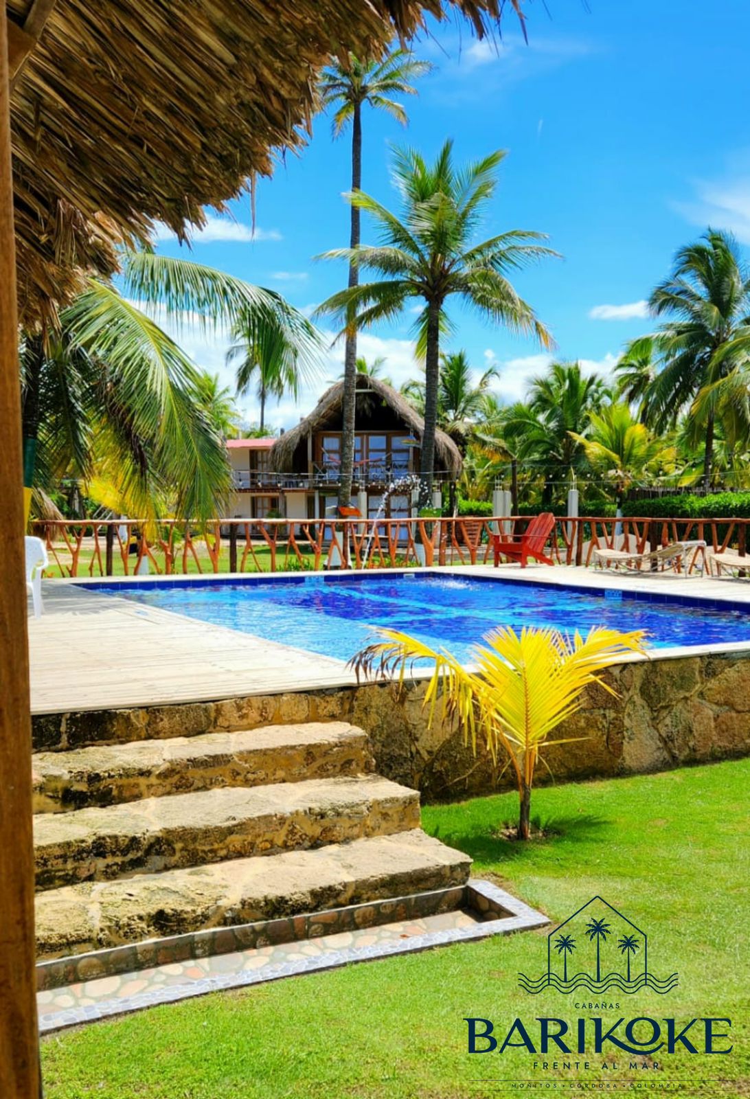 A clear blue swimming pool is bordered by a stone deck and vibrant green grass. Palm trees provide shade, and a thatched-roof structure is visible in the background. Bright seating options are arranged nearby, inviting relaxation in the tropical setting.