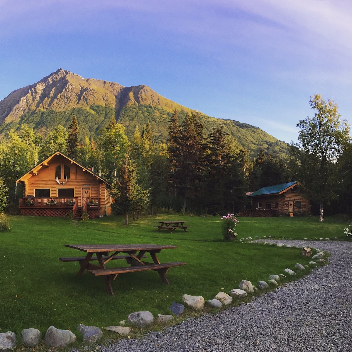 The exterior of the Caribou Cabin is surrounded by lush greenery, set against a mountain backdrop. A gravel pathway leads to the cabin, with picnic tables placed on the grassy area, inviting outdoor gatherings. The clear sky adds to the serene atmosphere.