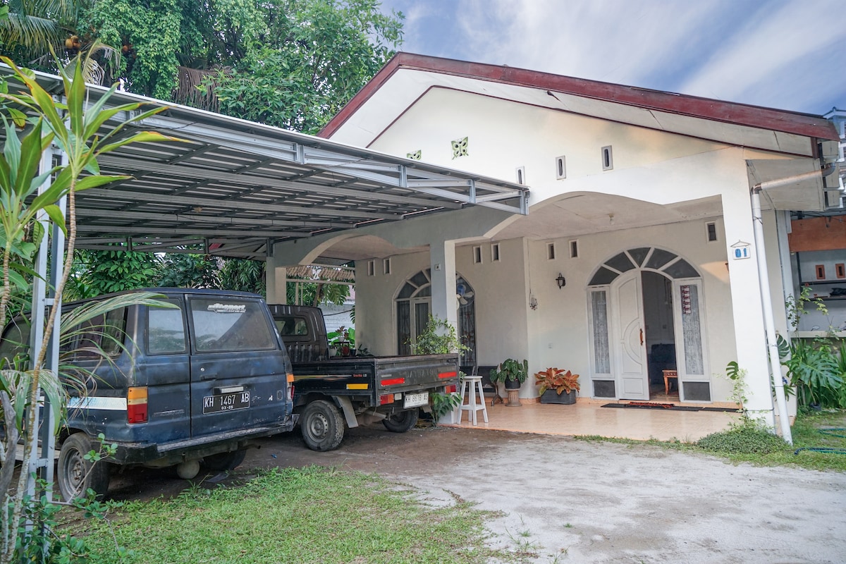 A carport is seen at the front of the rectangular house, which features a covered entrance with decorative archways. Two parked vehicles are visible, surrounded by greenery and potted plants, contributing to a suburban atmosphere.