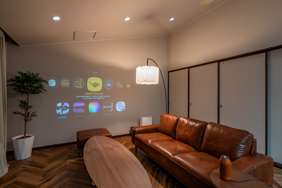 A comfortable living area features a brown leather sofa and a rounded wooden coffee table. A floor lamp provides soft lighting, while a projection on the wall displays various entertainment options. Additional greenery is added to the space with a potted plant.