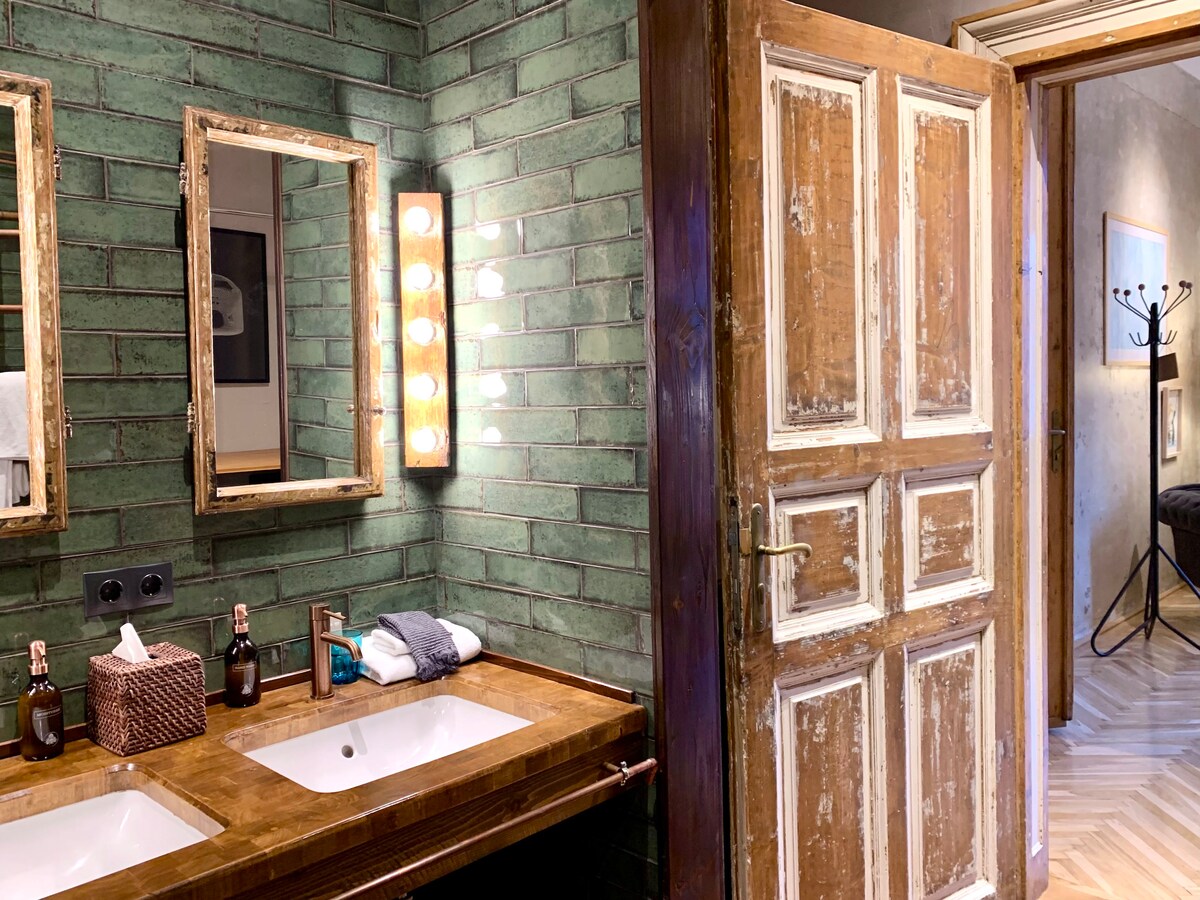 A bathroom features dual sinks set into a wooden countertop, with vintage mirrors above reflecting the green tiled walls. A rustic door adds character, while warm lighting creates a soft atmosphere. Towels and toiletries are arranged neatly on the countertop and in a woven basket.