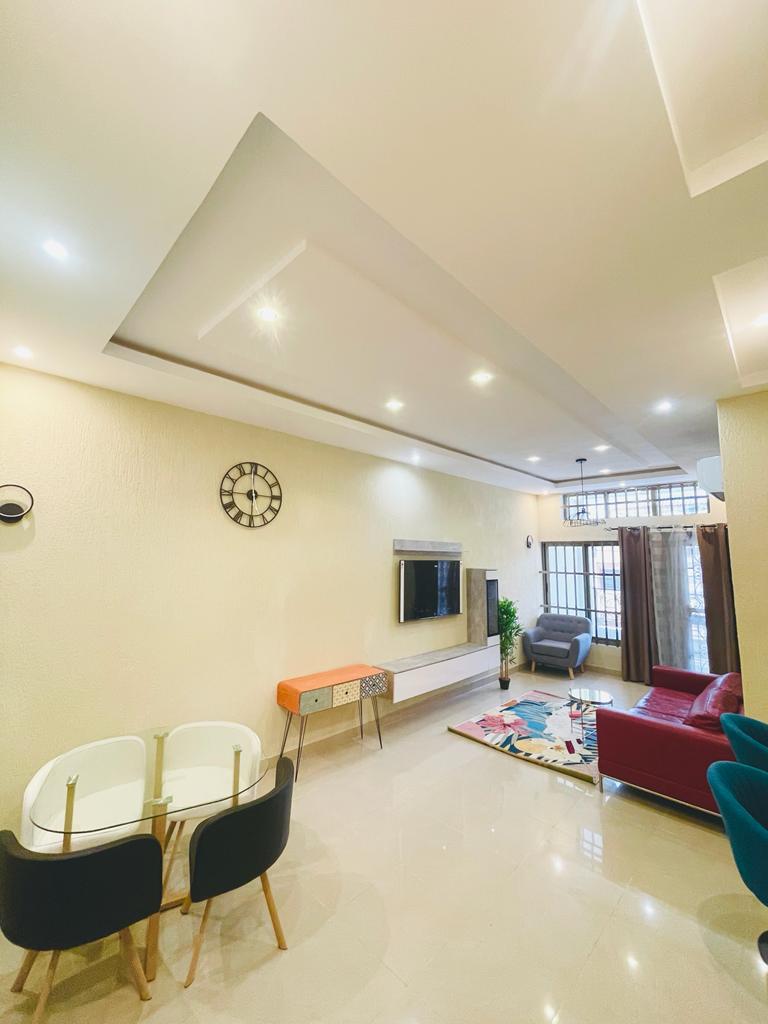 A modern living area features a flat-screen television mounted on the wall, with a stylish storage unit below. A round dining table is accompanied by four chairs, and a colorful rug adds visual interest to the floor. Large windows allow for natural light, enhancing the spaciousness.