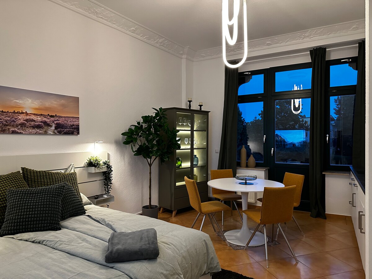 A modern studio layout is shown, featuring a spacious bed with neutral linens and decorative pillows. A glass-front cabinet displays decorative items and plants. A round dining table with four yellow chairs is positioned next to large windows, offering evening views and natural light.