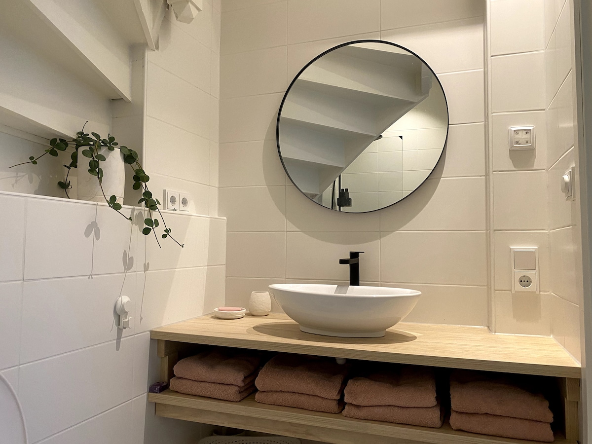 A modern bathroom is presented, featuring a round mirror above a stylish sink on a wooden countertop. Soft towels are neatly arranged on shelves below, while greenery adds a touch of nature. Clean, white tiles contribute to a bright and inviting space.