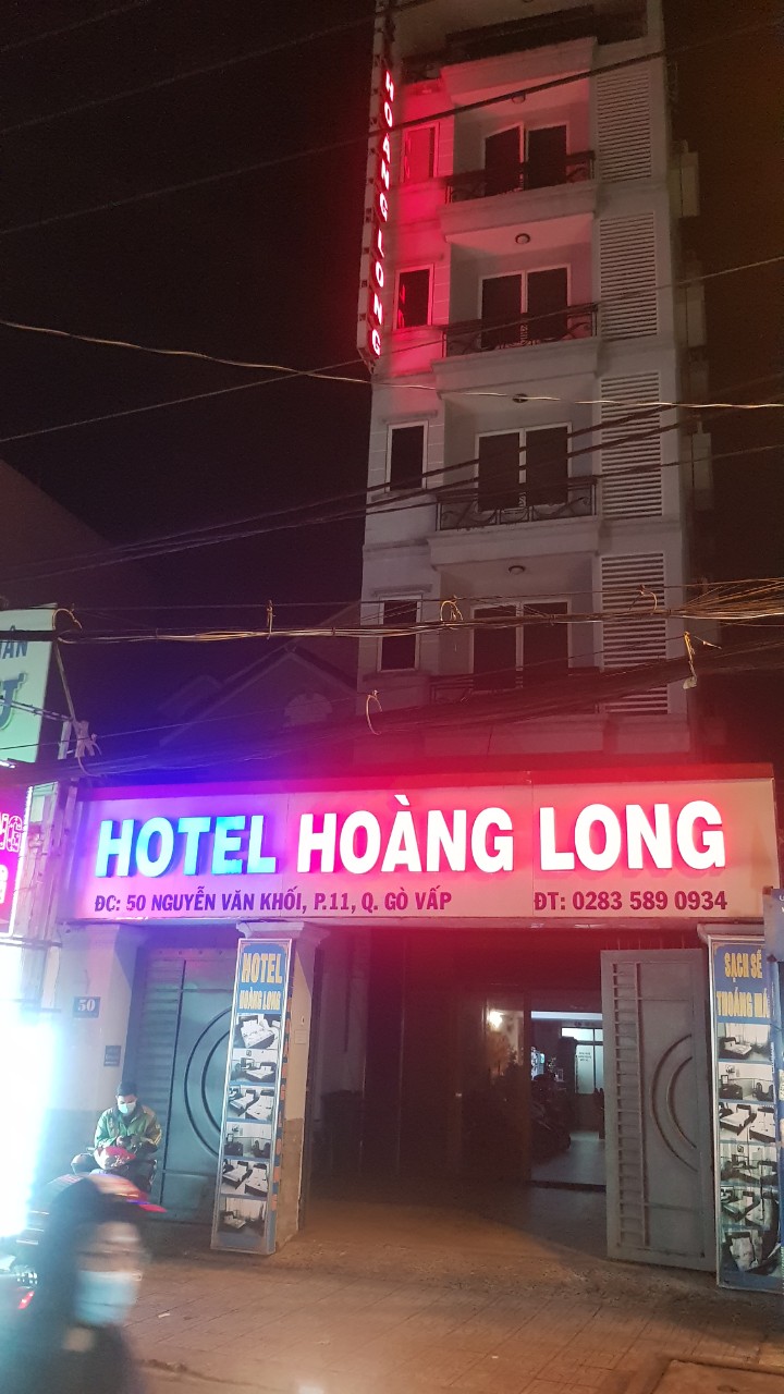 The exterior of Hotel Hoàng Long is illuminated by vibrant red and blue lights, emphasizing the hotel’s name. The multi-story building features a simple façade with a welcoming entrance. Surrounding elements such as signage and street details contribute to the lively atmosphere.