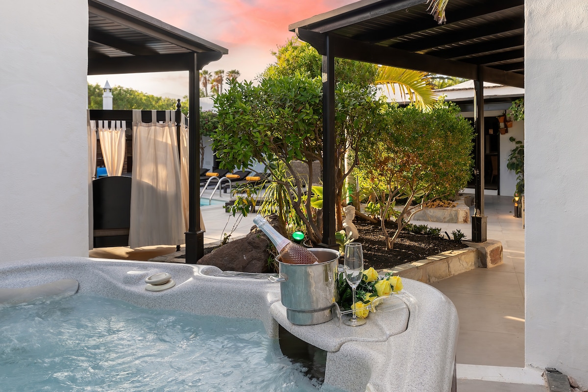 A private jacuzzi is positioned beside lush greenery and bright yellow flowers, with a bottle of champagne cooling in an ice bucket. In the background, a glimpse of the heated swimming pool and outdoor lounge area is visible under a covered pergola.