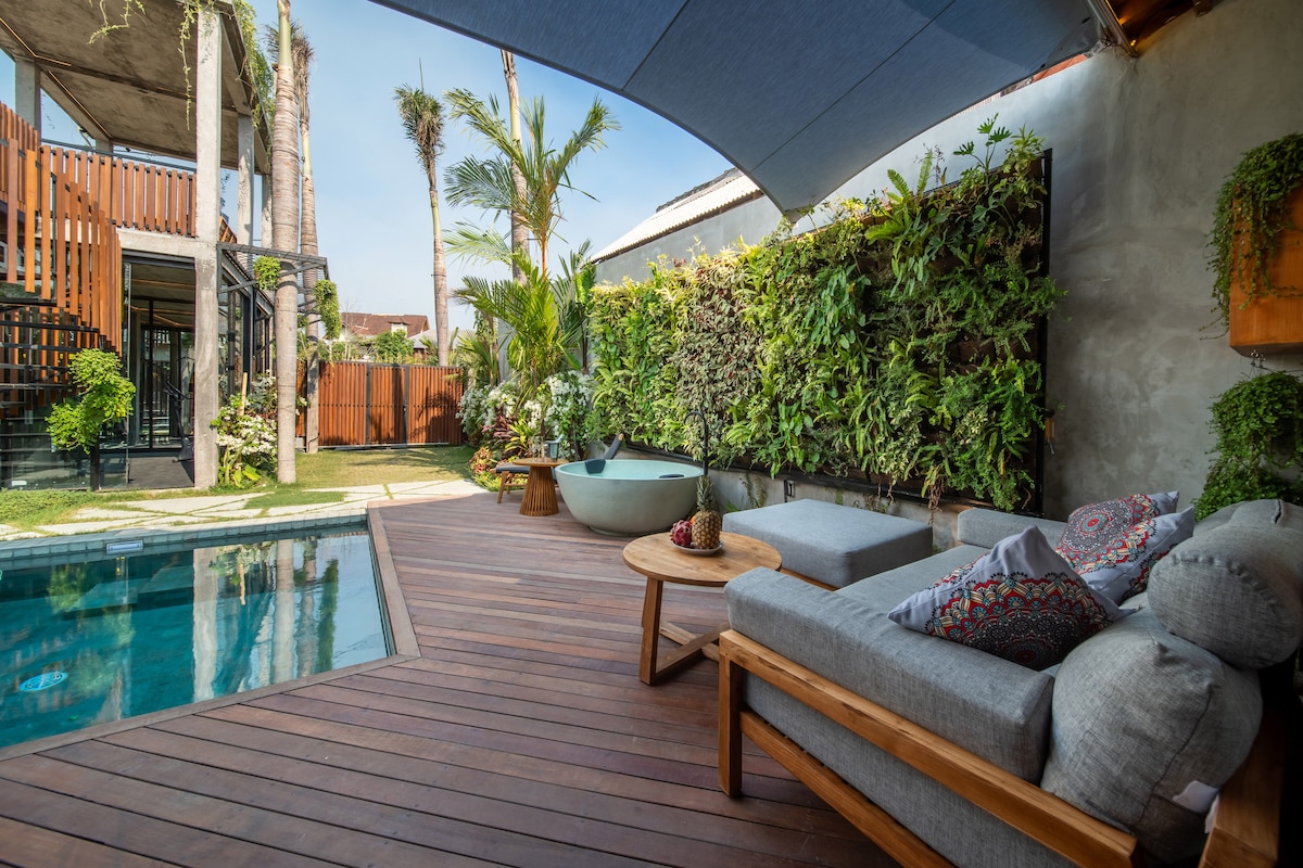 A spacious outdoor area features a wooden deck beside a refreshing swimming pool. Comfortable seating is arranged near a jacuzzi, surrounded by vibrant greenery. Tall palm trees and lush plants create a serene ambience, while a shade sail offers relief from the sun.