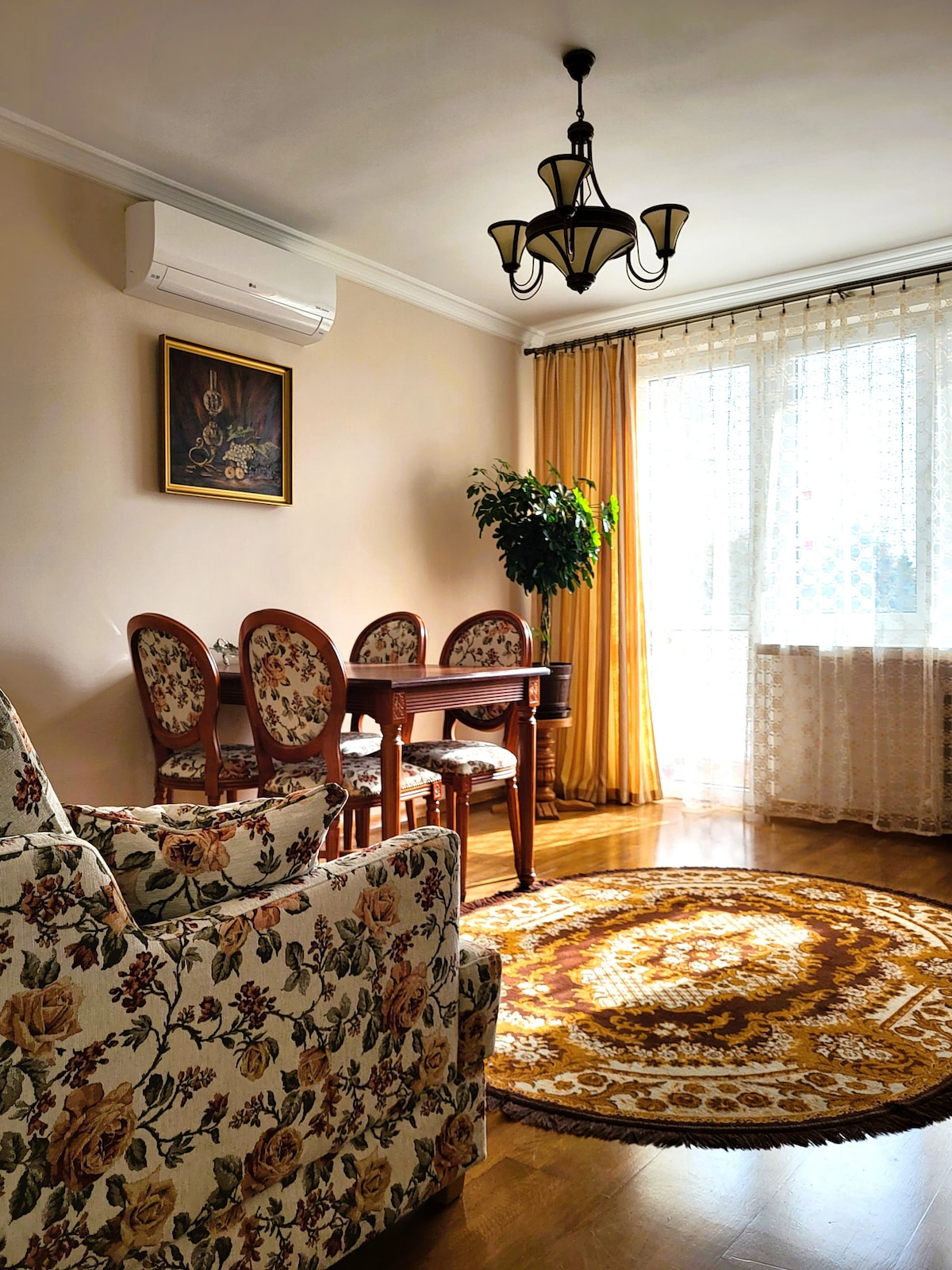 A bright living area is furnished with a floral-patterned armchair and a dining set for four. Natural light filters through sheer curtains, casting soft shadows on the wooden floor. A circular rug in warm tones enhances the inviting ambiance, while an air conditioning unit maintains comfort.