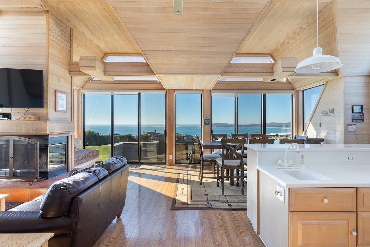 The open Great Room features large windows framing the water views. A comfortable seating area with a dark leather sofa is positioned near a fireplace. A dining table with wooden chairs is visible adjacent to the fully equipped kitchen, highlighting a seamless flow between spaces.