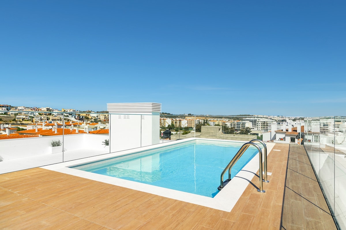 A rooftop swimming pool is depicted against a clear blue sky, offering a view of the surrounding city and countryside. The pool area is surrounded by a wooden deck, with a sleek railing visible along the edge, creating a modern and inviting space.