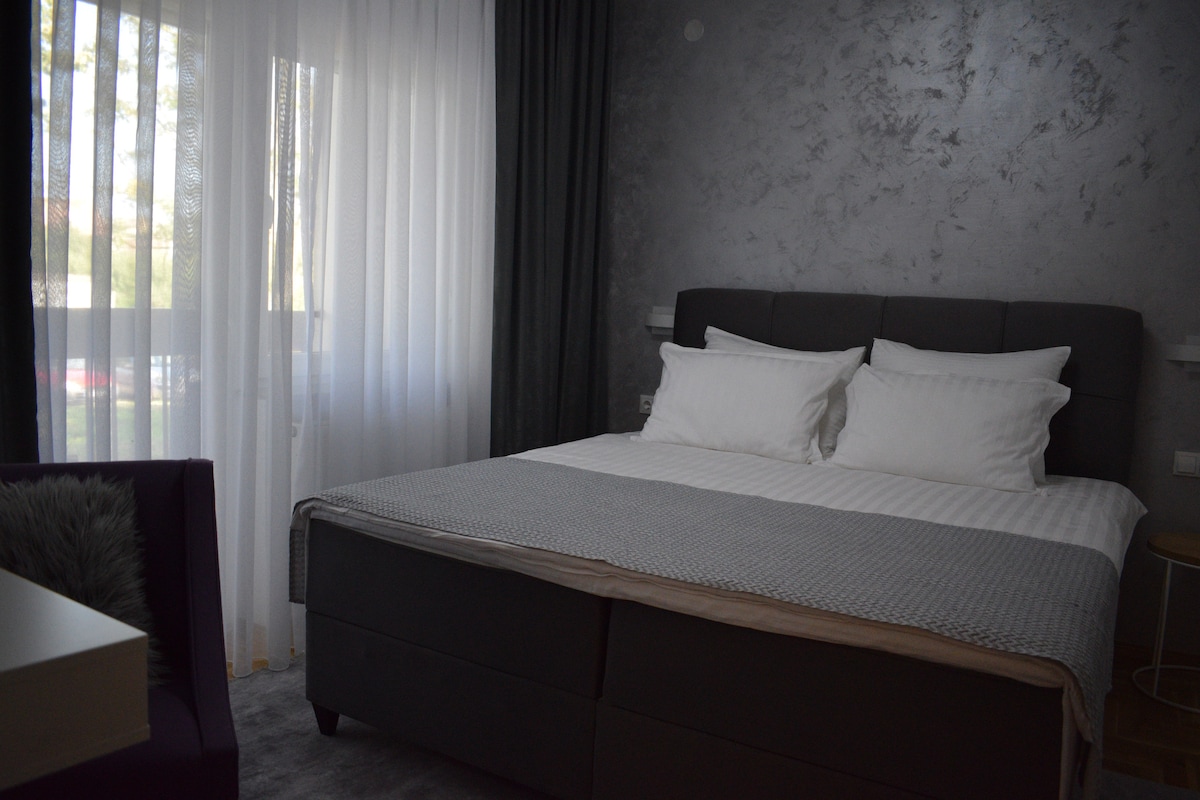 A modern bedroom is seen with a spacious bed featuring neutral-colored bedding and two pillows. Sheer curtains allow natural light to filter in, adding a soft glow. A small bedside table is positioned next to the bed, with a cozy chair nearby.
