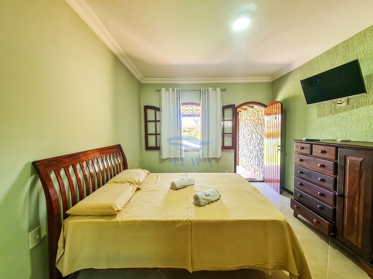 A cozy bedroom is furnished with a spacious bed covered in yellow linens. Two large windows are adorned with white curtains, allowing natural light to illuminate the room. A wooden dresser stands against one wall, while a TV is mounted nearby, contributing to a comfortable atmosphere.